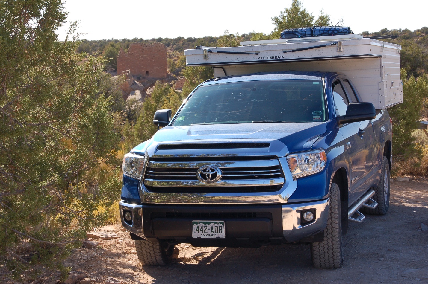 A truck is parked in front of a blue car on a dirt road

Description automatically generated